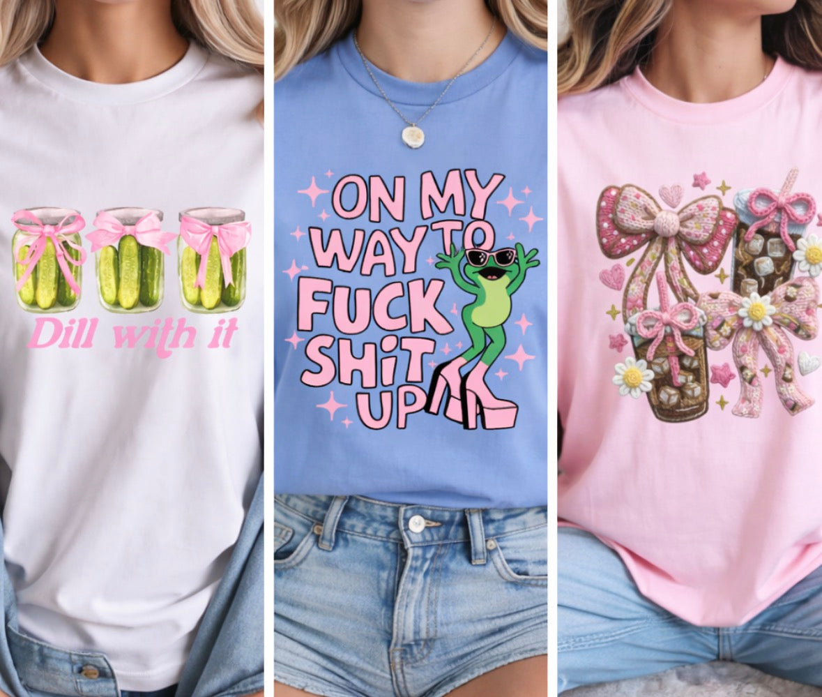 Women wearing a white “Dill With It” pickle shirt, a blue “On My Way to Fuck Shit Up” frog graphic tee, and a pink coquette bow iced coffee tshirt.