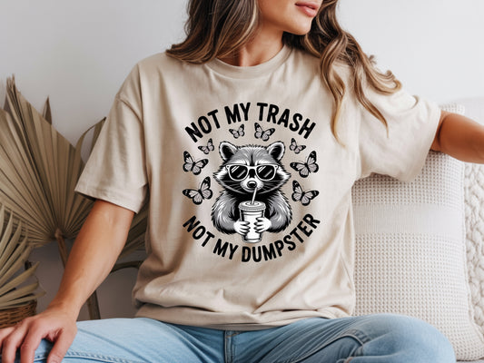 Beige raccoon shirt in oversized unisex fit featuring “Not My Trash Not My Dumpster” quote with a raccoon and butterflies. 