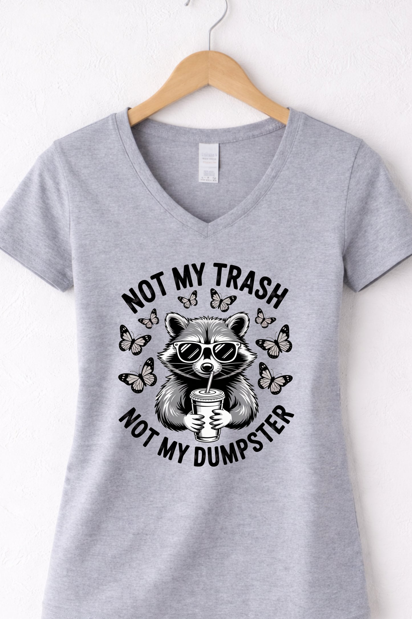 Grey women’s v-neck raccoon shirt hanging display featuring “Not My Trash Not My Dumpster” quote.