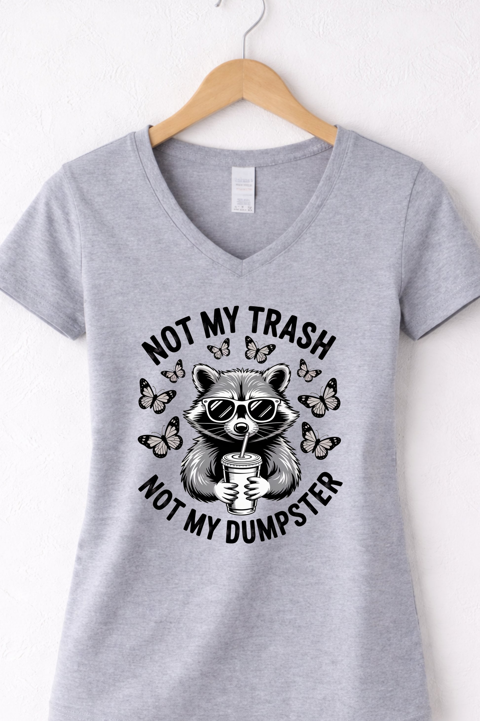 Grey women’s v-neck raccoon shirt hanging display featuring “Not My Trash Not My Dumpster” quote.