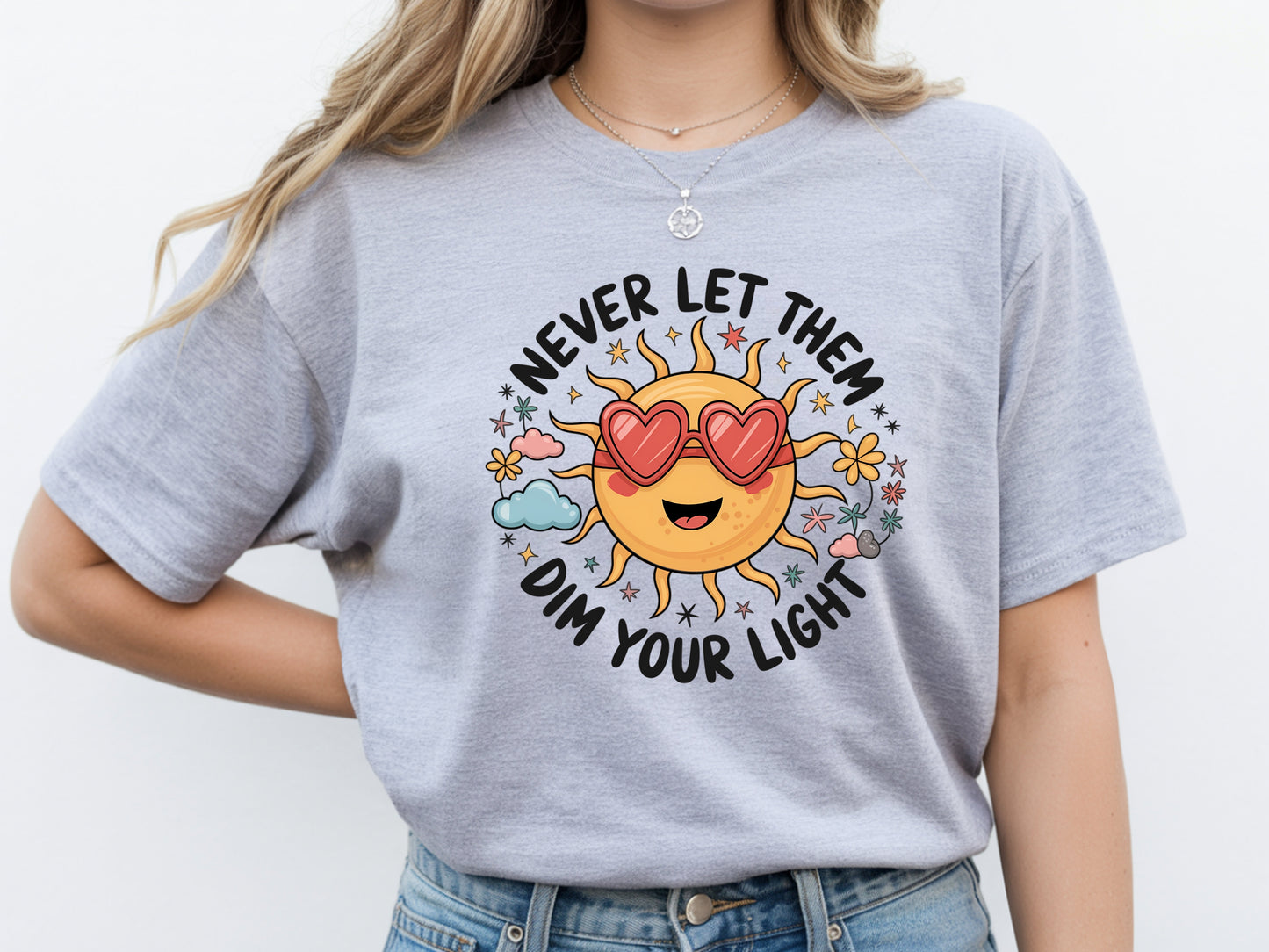 Never let them dim your light shirt with a cute sun graphic and "Never let them dim your light" text on a grey unisex shirt.