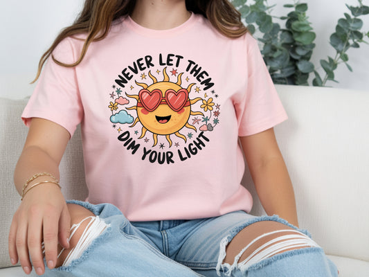 Never let them dim your light shirt with a cute sun graphic and "Never let them dim your light" text on a pink unisex shirt.