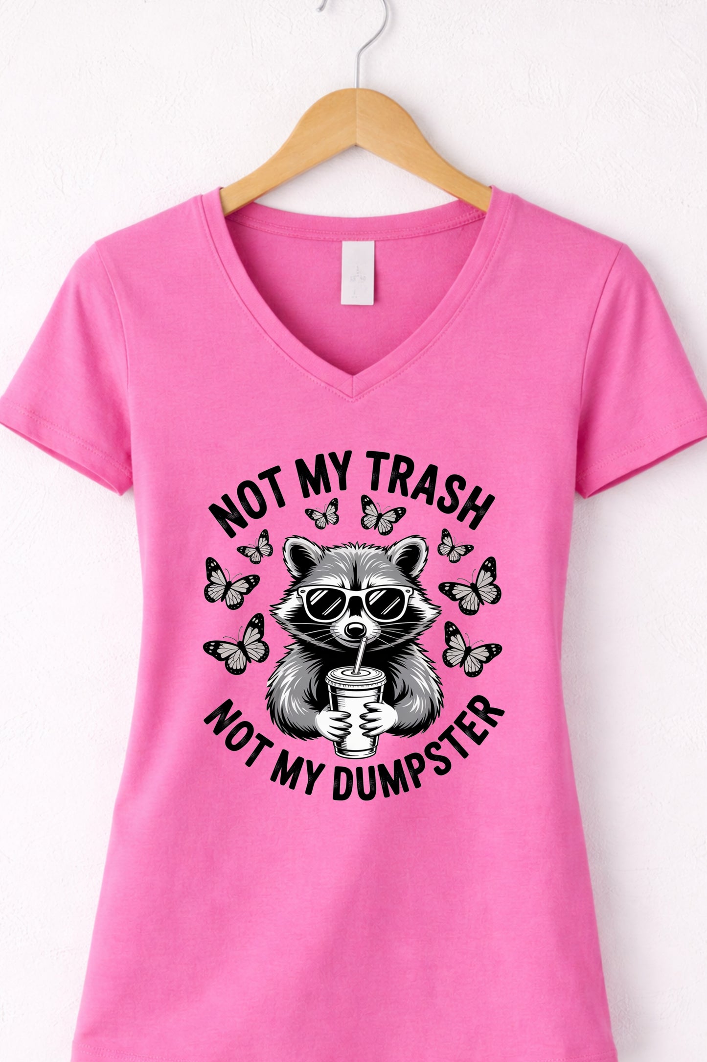 Pink women’s v-neck raccoon shirt hanging display featuring “Not My Trash Not My Dumpster” quote.