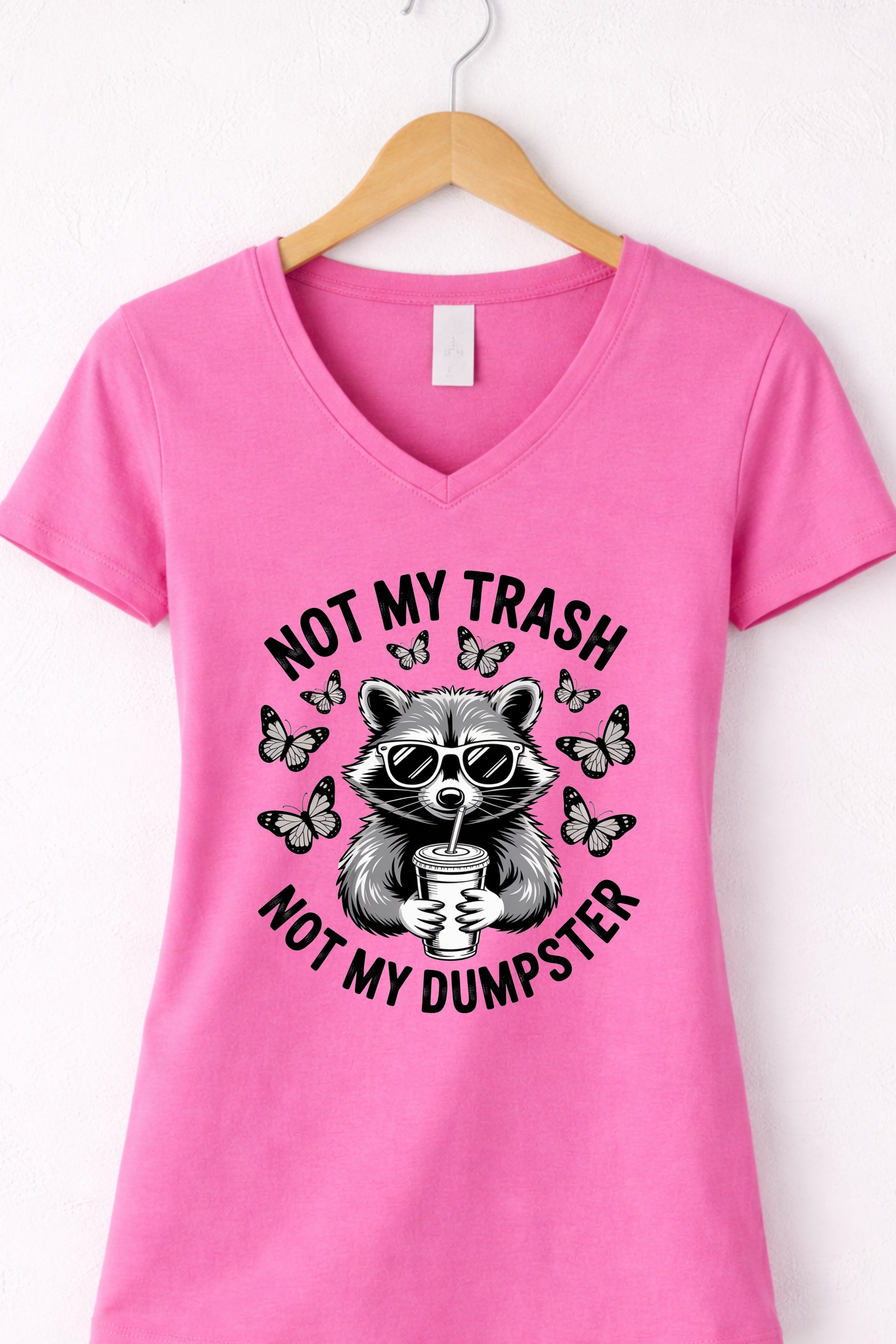 Pink women’s v-neck raccoon shirt hanging display featuring “Not My Trash Not My Dumpster” quote.